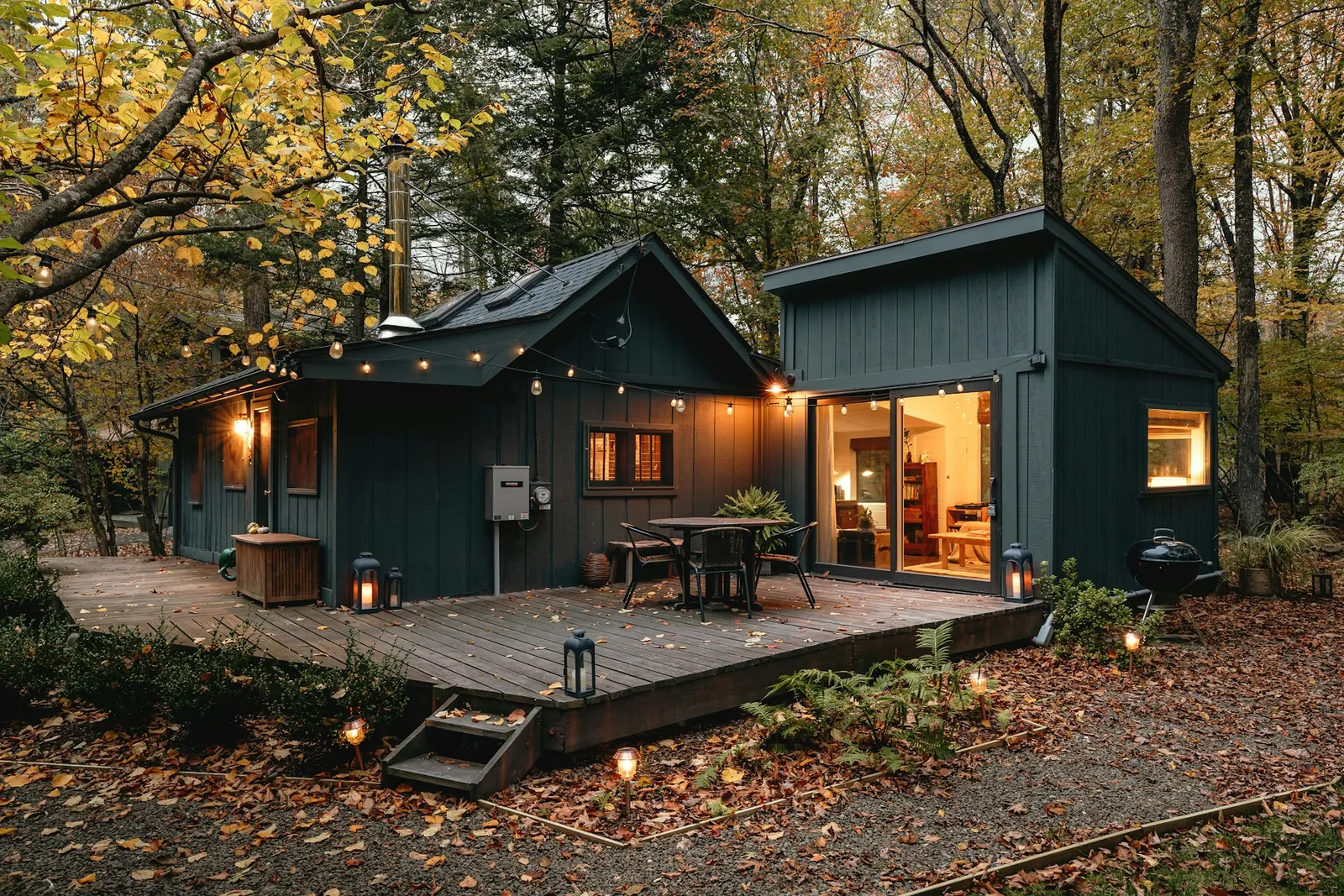 A dark green cabin at dusk in an autumn forest, string lights glowing over a wooden deck with a bistro table and lanterns, warm light spilling from the open glass doors