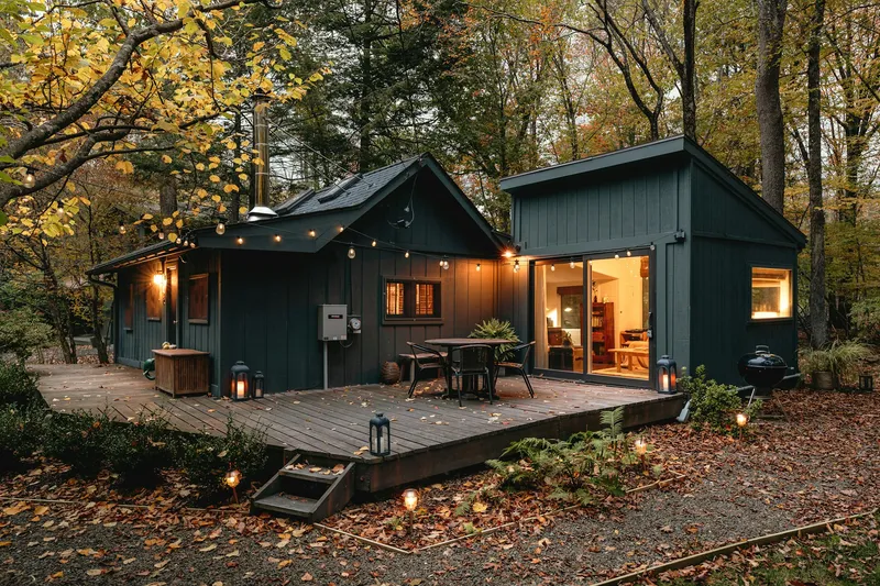 A dark green cabin at dusk in an autumn forest, string lights glowing over a wooden deck with a bistro table and lanterns, warm light spilling from the open glass doors