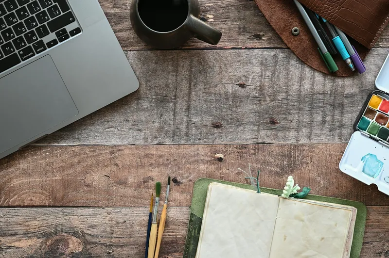 Open laptop next to a notebook, diary, and a cup of tea on a wooden desk, viewed from above