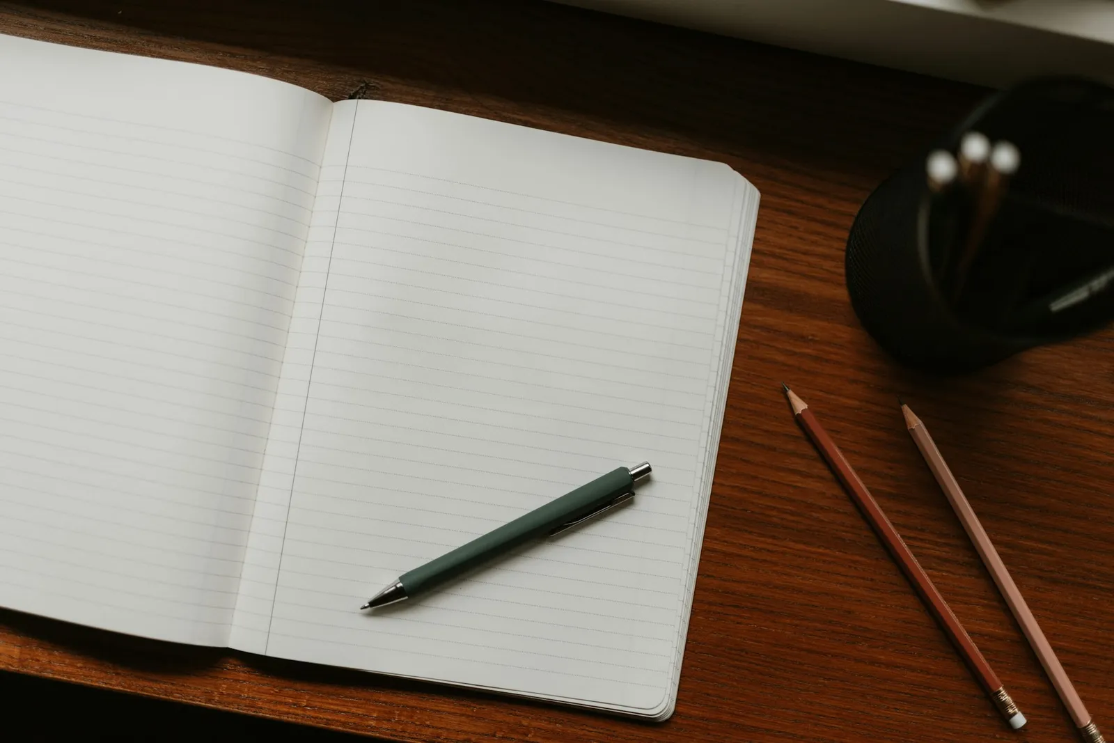 Open spiral notebook with blank lined pages on a wooden desk next to a black pen and a pair of pencils