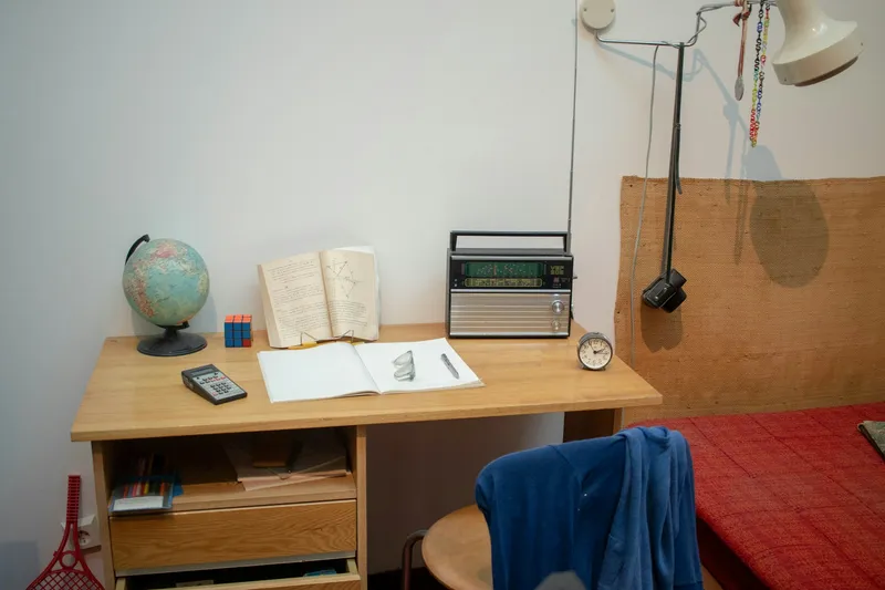 Vintage student desk with a small globe, open book, calculator, notebook with pen, retro radio, alarm clock, and a chair with a blue sweater beside a red-blanketed bed