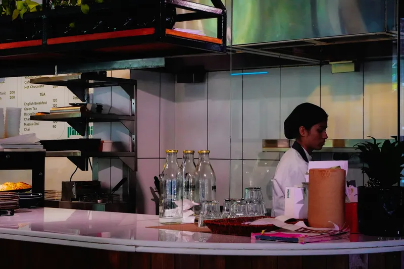 Person in apron working behind a wooden cafe counter preparing drinks in warm light, face not visible