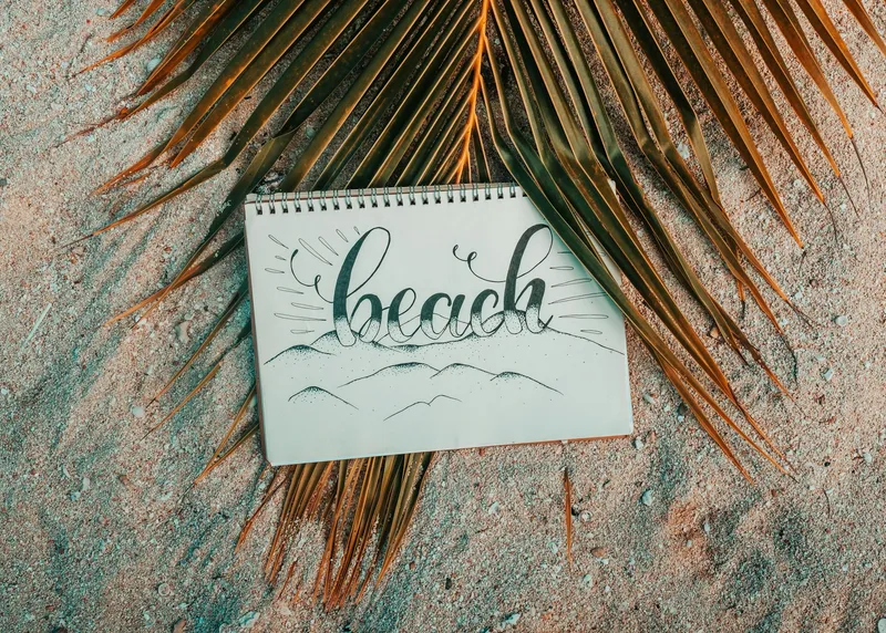 Overhead flat-lay of an open sketchpad and pen resting on light beach sand